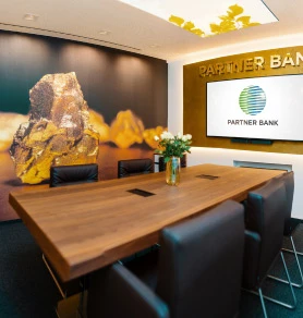 Ein moderner Besprechungsraum mit einem großen Holztisch, einem goldenen Balken an der Wand und dem Logo der Partner Bank auf einem Bildschirm.