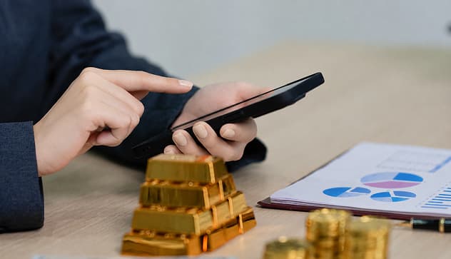 Person using a smartphone while analyzing gold bars and financial charts on a desk, representing investment decisions during uncertain economic times.