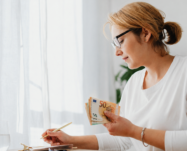 Frau verwaltet ihre Finanzen zu Hause, berechnet Ausgaben und hält Bargeld in der Hand, als Symbol für Herausforderungen von Frauen beim Investieren in Aktien und beim Aufbau finanzieller Sicherheit.